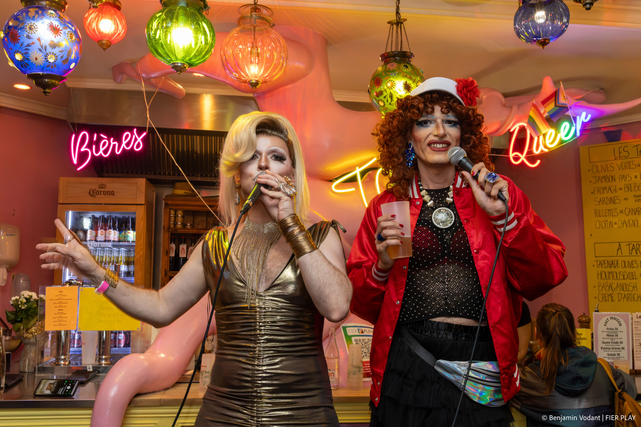 Deux drag queens avec micros dans un bar décoré “Queer” et lanternes colorées