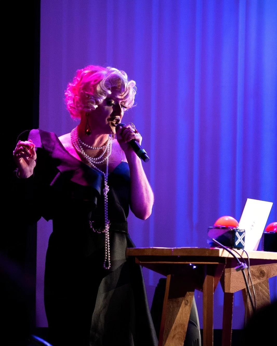 Drag queen en robe noire, perruque blonde et collier de perles animant un quiz sur scène avec micro et lumière violette
